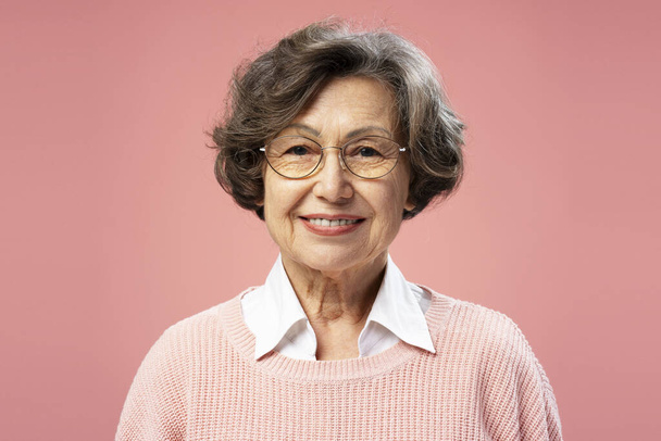 Усміхаючись красива старша жінка, бабуся, одягнена в окуляри, дивиться на камеру, що стоїть ізольовано на рожевому фоні. Концепція реклами - Фото, зображення