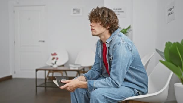 Ένας νεαρός Ισπανός με σγουρά μαλλιά και τζιν σακάκι κάθεται σε ένα λευκό δωμάτιο, κοιτάζοντας ένα ανοιχτό διαβατήριο.. - Πλάνα, βίντεο
