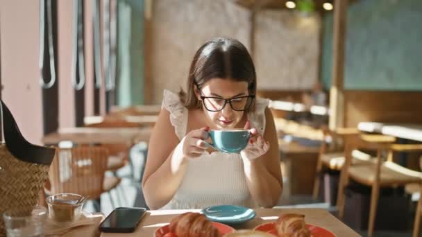Ηλιοφωτισμένο πορτραίτο μιας όμορφης, νεαρής Ισπανίδας με γυαλιά σε ένα καφέ, που χύνει γλυκά ζάχαρη στον καυτό πρωινό καφέ της στο τραπέζι του πρωινού - Πλάνα, βίντεο