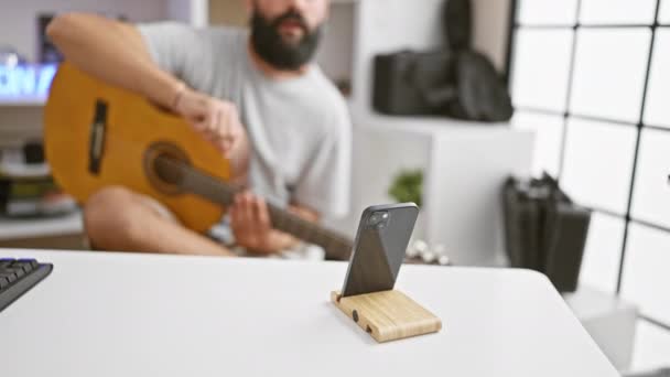 Smiling bearded man playing acoustic guitar indoors, recording with smartphone on wooden stand. - Footage, Video