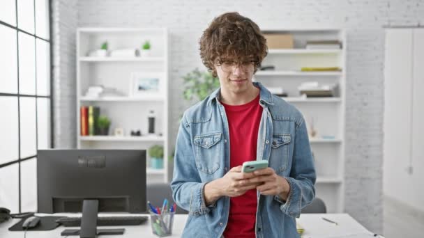Un jeune homme élégant avec des cheveux bouclés et des lunettes textes sur un smartphone dans un cadre de bureau moderne. - Séquence, vidéo