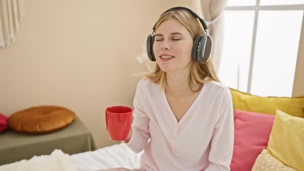 Een jonge blanke vrouw geniet van muziek met koptelefoon en koffie in een gezellige slaapkamer ochtend. - Video