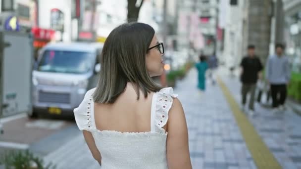 Rückansicht einer schönen hispanischen Frau, die die Tokyostraße entlanggeht, durch ihre Brille auf die städtische Umgebung blickt und das Leben und die architektonische Schönheit der Stadt einfängt - Filmmaterial, Video