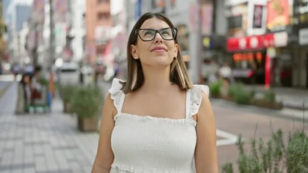 Mooie Latijns-Amerikaanse vrouw in bril, nonchalant staande en vrolijk glimlachend, poserend haar vertrouwen in tokyo 's moderne straten, rondkijkend, genietend van Japans stadsgezicht. - Video