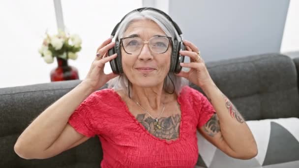 Fiduciosa donna di mezza età che si toglie gioiosamente le cuffie, rilassarsi dopo aver goduto della musica sul suo divano a casa - Filmati, video