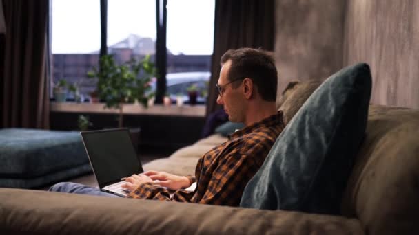Relaxed serious Caucasian guy student freelancer using laptop device leaning on sofa at home office, focused entrepreneur working distantly typing on notebook in apartment - Footage, Video