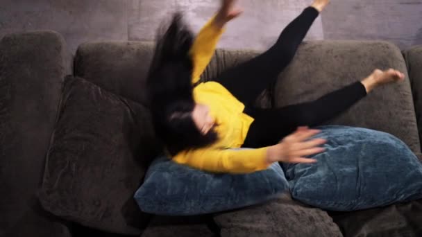 Happy positive Asian woman fallen on cozy couch, enjoying lazy weekend relaxation time alone at home. Smiling young female resting on sofa, raising arms stretching muscles, reposing indoors. - Footage, Video