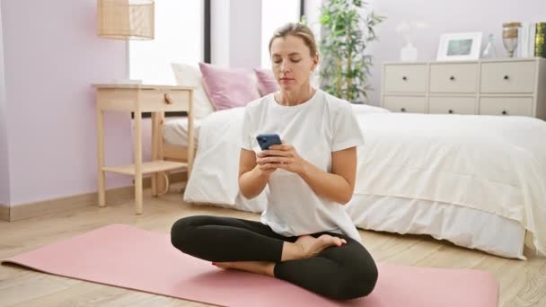 Een serene jonge blonde vrouw mediteert op een roze yoga mat in haar slaapkamer, belichaamt welzijn en evenwicht. - Video