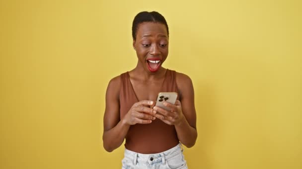 Emocionada mujer afroamericana, orgullosamente celebrando la victoria y el éxito, levanta el brazo mientras envía mensajes de texto en su teléfono amarillo contra un fondo aislado - Metraje, vídeo