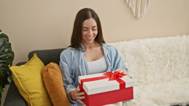 Joie exténuante, confiante, jeune femme hispanique souriante tandis que assise sur son confortable canapé à la maison, déballer son cadeau d'anniversaire surprenant - Séquence, vidéo