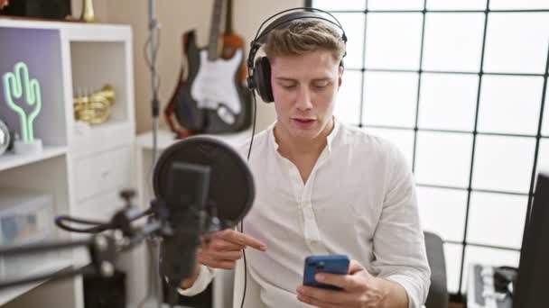 Впевнений, молодий кавказький чоловік є багатостороннім радіорепортером, розмиваючи гарячі новини через мікрофон, читаючи зі свого екрану смартфона на етапі трансляції в приміщенні радіостудії - Кадри, відео