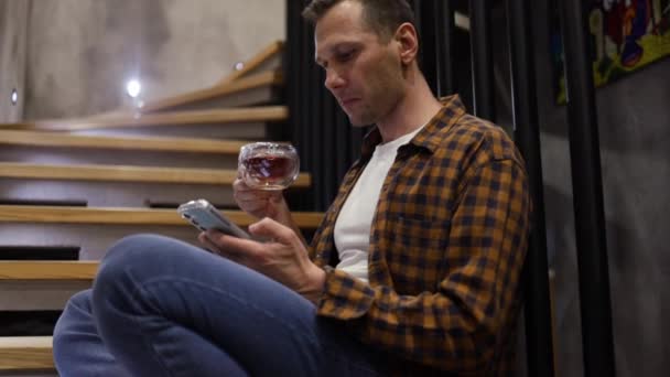 Biały mężczyzna siedzi na schodach w nowoczesnym mieszkaniu, pijąc poranną kawę, przewijając wiadomości przez telefon. Biznes człowiek za pomocą smartfona w swoim biurze. - Materiał filmowy, wideo