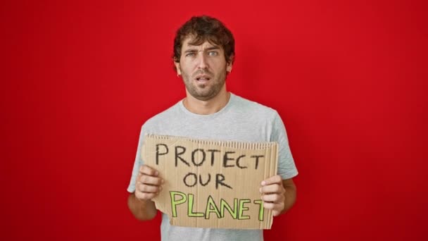 Ο μπερδεμένος νεαρός που κρατά το πανό "προστατέψτε τον πλανήτη μας" εκπέμπει σκεπτικιστικό βλέμμα με σαρκαστική έκφραση με ανοιχτό στόμα σε κόκκινο απομονωμένο φόντο - Πλάνα, βίντεο