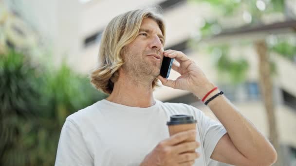 Красивый мужчина с длинными волосами разговаривает по телефону, держа кофе на городской улице - Кадры, видео