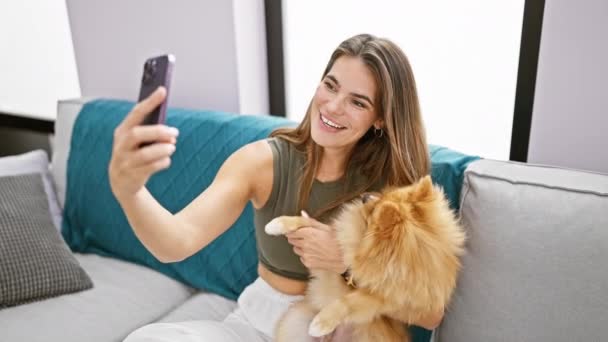 Ontspannen jonge Spaanse vrouw genieten van een grappige video oproep op haar mobiel met haar gelukkige hond aan haar zijde, zittend op de woonkamer bank thuis. - Video