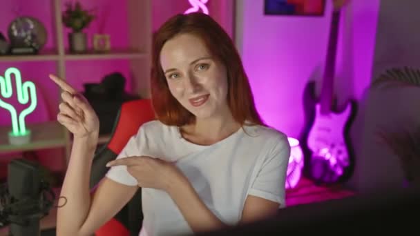 陽気な若い赤毛の慎重な女性ストリーマーは,カメラを見て,笑顔と自宅のオフィスの夜にゲーム室で指摘 - 映像、動画