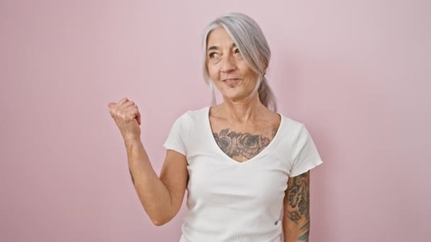 Уверенная женщина среднего возраста с седыми волосами стоит бодро, показывая большие пальцы вверх, широко улыбаясь, сияя оптимизмом на изолированном розовом фоне - Кадры, видео