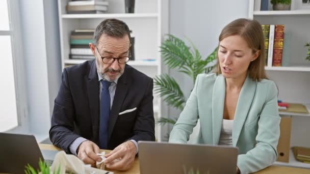 Zwei Geschäftsleute niesen im Büro zusammen, während sie hart an einem Laptop arbeiten - ein klarer Ausdruck für eine Grippeerkrankung im Büro. - Filmmaterial, Video