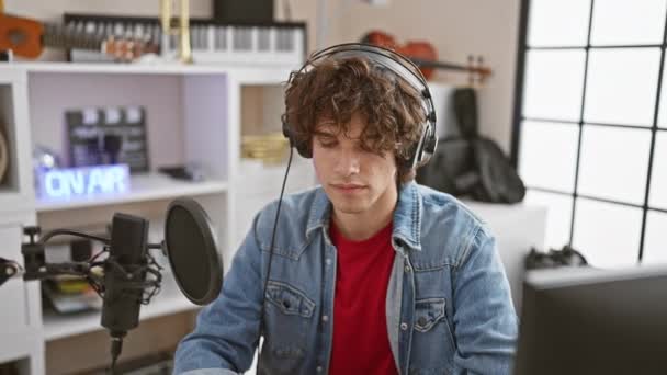 A young man with curly hair and a denim jacket records vocals while wearing headphones in a modern home music studio. - Footage, Video