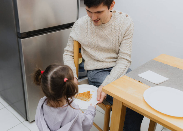 Una piccola ragazza caucasica serve un pezzo di galette reale in un piatto a un ragazzo adulto seduto a un tavolo in cucina vicino al frigorifero, vista laterale da vicino. - Foto, immagini