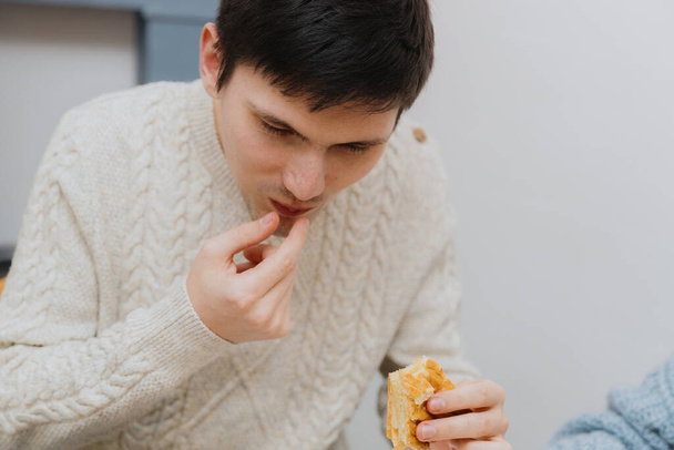 One young handsome Caucasian guy cheerfully takes out a surprise gift from a piece of royal galette from his mouth, sitting at a table in the kitchen near the refrigerator, close-up side view. - Photo, Image