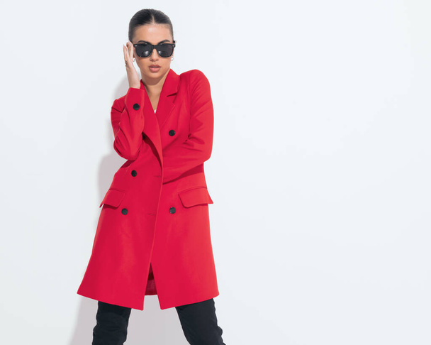 портрет привлекательной прохладной женщины в красном пальто, фиксирующей солнцезащитные очки и уверенной в себе, позируя на белом фоне - Фото, изображение