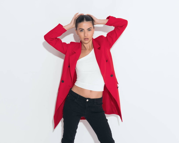 bella donna sexy in cappotto rosso che tiene le braccia in su e posa con le mani dietro la testa in modo sicuro su sfondo bianco - Foto, immagini