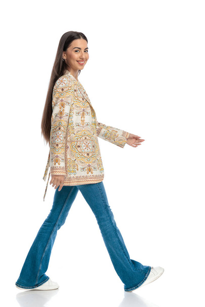 side view picture of beautiful casual girl in jacket with blue jeans walking and smiling in front of white background - Photo, image