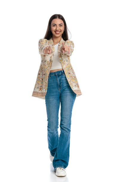 full length picture of beautiful young woman in blue jeans walking, pointing fingers forward and smiling in front of white background - Photo, Image