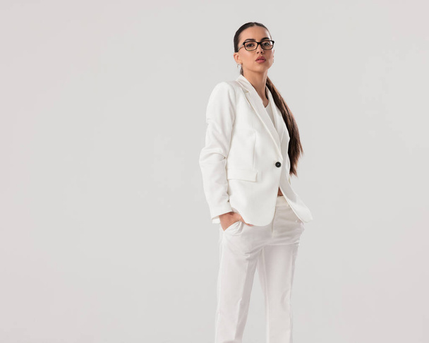 уверенная молодая деловая женщина в белом костюме в очках, держась за руки в карманах и позируя на сером фоне - Фото, изображение