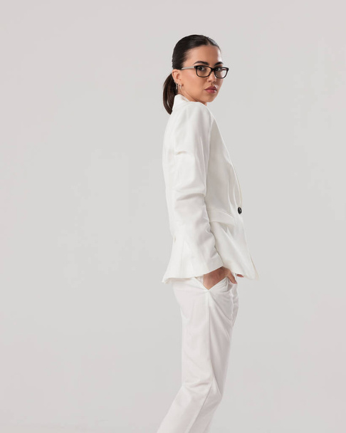 side view of elegant fashion woman with glasses holding hands in pockets and looking forward in front of grey background - Photo, Image