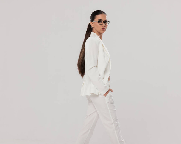 vista laterale della bella giovane donna d'affari che si tiene per mano in abito bianco, indossando occhiali e calpestando lo sfondo grigio - Foto, immagini