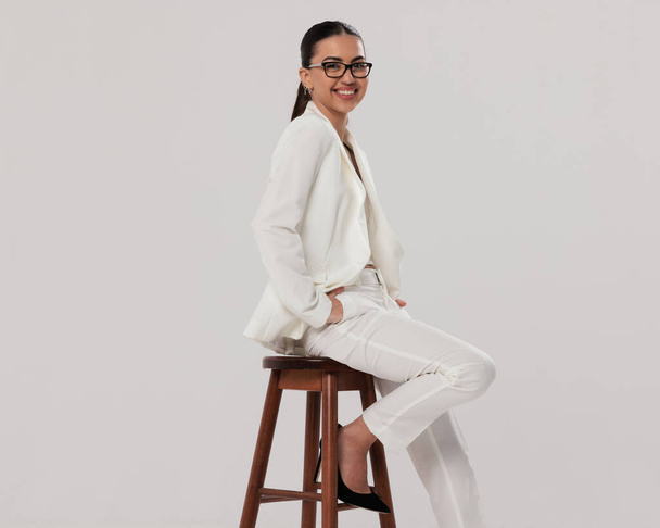 happy businesswoman with glasses in white suit holding hands in pockets and laughing while sitting on wooden chair in front of grey background - Photo, Image
