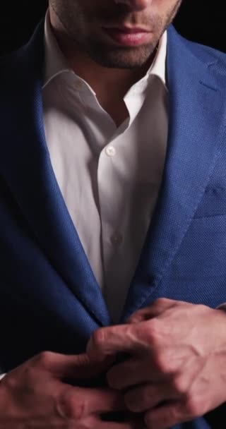 elegante jonge zakenman in blauw pak naar beneden kijkend, openingspak, vooruit kijkend terwijl hij poseert voor een zwarte achtergrond - Video