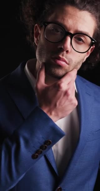 hombre elegante guapo con gafas sensualmente tocando la cara, mirando hacia adelante mientras la cámara se acerca en el fondo negro - Imágenes, Vídeo