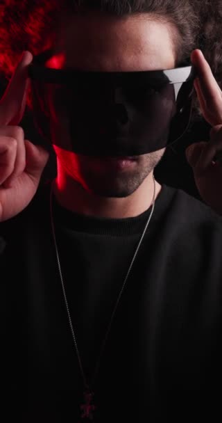 pionowe wideo przypadkowy młody człowiek z kręconymi włosami dostosowując futurystyczne okulary przeciwsłoneczne i naszyjnik patrząc do przodu na czarnym tle - Materiał filmowy, wideo
