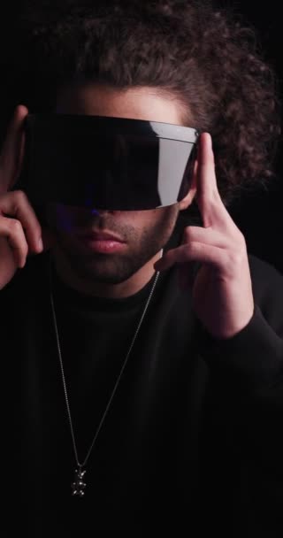 κοντινό βίντεο με σέξι νεαρό άνδρα με μαύρη κουκούλα να προσαρμόζει φουτουριστικά γυαλιά ηλίου, να σταυρώνει τα χέρια και να κοιτάει στο πλάι σε μαύρο φόντο - Πλάνα, βίντεο