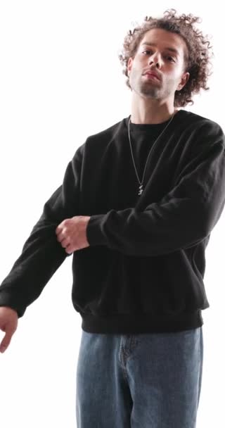 sexy hombre casual con pelo rizado ajustando las mangas y el cuello de la sudadera mientras que es fresco y posando de una manera segura sobre fondo gris - Imágenes, Vídeo