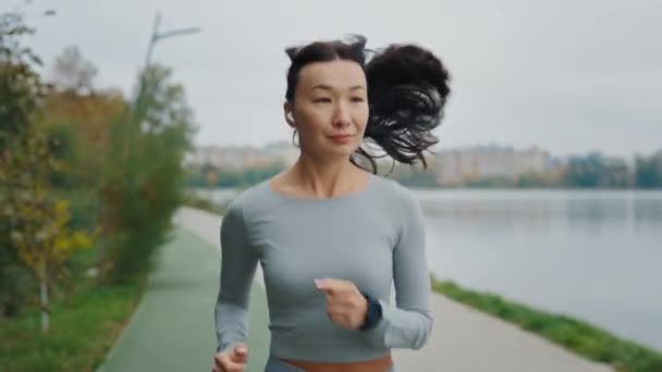 Asijské ženské běžec nosit sluchátka poslouchat hudbu je jogging poblíž města místní jezero. Sportovní sportovkyně ve sluchátkách běžící na místním jezeře. Školení žen venku - Záběry, video