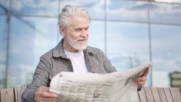Maschio di età positiva con la barba godendo di leggere giornale in luogo urbano mentre seduto sulla panchina. Ritratto di persona anziana elegante attentamente ricerca articolo nella stampa del mattino con bel tempo. - Filmati, video