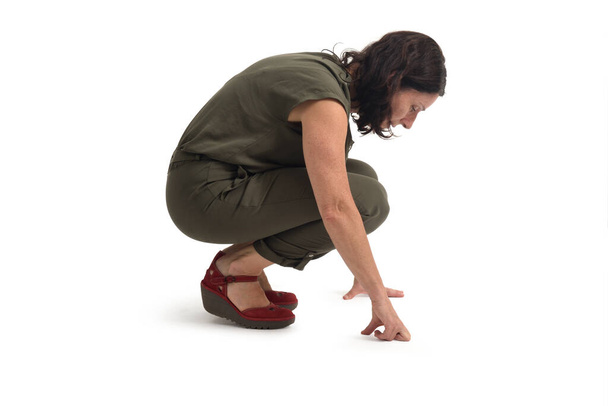 vista lateral de una mujer en cuclillas buscando o mirando algo en el suelo sobre fondo blanco - Foto, Imagen