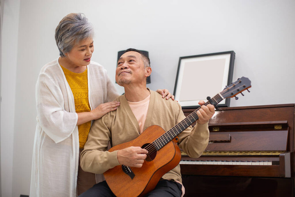 Ευτυχισμένο ζευγάρι ηλικιωμένων Ασιατών, χαλαρώστε στις διακοπές συνταξιοδότησης παίζοντας κιθάρα διασκεδάζοντας στο σαλόνι στο σπίτι, τραγουδώντας μαζί την Ευτυχία. Φροντίζει ο ένας τον άλλον στην υγεία σε μεγάλη ηλικία. - Φωτογραφία, εικόνα