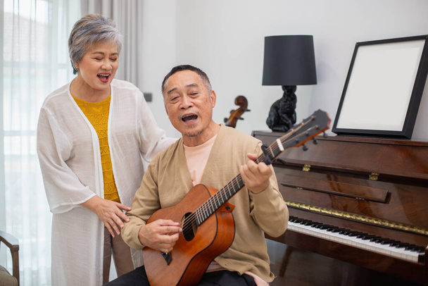 Glückliches asiatisches Senioren-Ehepaar lässt es sich im Rentenalter gemütlich machen, Gitarre spielen, im heimischen Wohnzimmer Spaß haben und gemeinsam singen. Fürsorge füreinander Gesundheit im Alter. - Foto, Bild