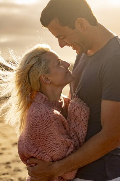 Люди в любові і золотий захід сонця на відкритому повітрі - чоловік і жінка пара обіймаються і виглядають один з одним з пляжем на задньому плані концепція захисту і життя разом назавжди - Фото, зображення