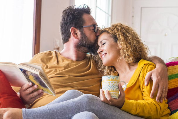 Glückliche schöne kaukasische Paare zu Hause genießen morgendliche Freizeitaktivitäten in entspannter Gemeinschaft - Liebes- und Beziehungsleben mit jungen Menschen, die auf dem bunten Sofa mit Buch und Kaffee sitzen - Foto, Bild