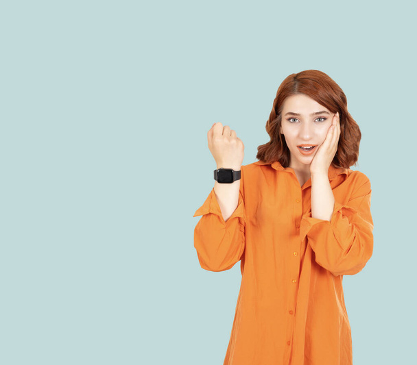 Akıllı saat ekranı gösteren genç, şaşkın kadın akıllı saat ekranı gösteriyor. Yanağına dokunan kızıl saçlı kızın portresi, açık mavi arka plan, fotokopi alanı. Teknoloji konsepti. - Fotoğraf, Görsel