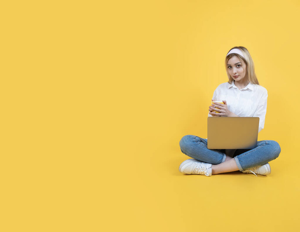 座っている若い女性はノートパソコンでコーヒーを飲んでいます. 全長のボディビュー 白人ブロンドの女の子の学生は足を横切りました. 黄色の背景が隔離されている. スペースをコピーする。 教育コンセプト. - 写真・画像