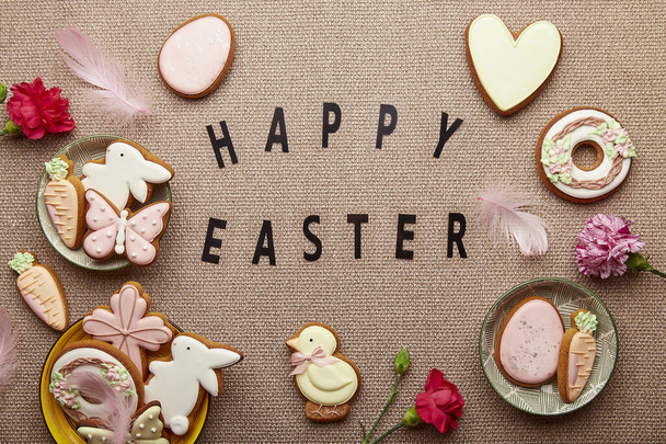 ハッピーイースターのテキストと春のパステル背景. 装飾されたイースタークッキー,ピンクの花,羽,卵の平らな層 - 写真・画像