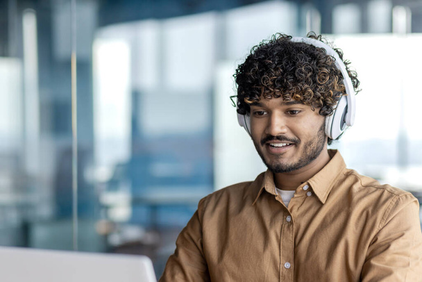 Молодой индийский программист, работающий в офисе с ноутбуком, человек в наушниках, кодирующий новое программное обеспечение, бизнесмен-разработчик доволен достигнутыми результатами улыбаясь. - Фото, изображение