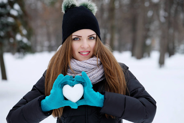 Egy tinédzser lány téli kalapban, kabátban és kesztyűben egy téli parkban egy hóból készült szívet tart a kezében. - Fotó, kép